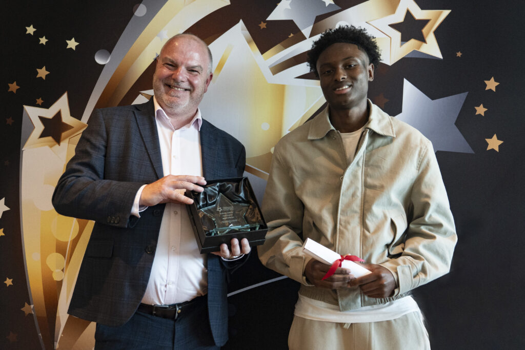 A smiling man in a suit holds a boxed award, standing next to a smiling young man in a beige jacket holding a rolled-up certificate, against a star-patterned background.