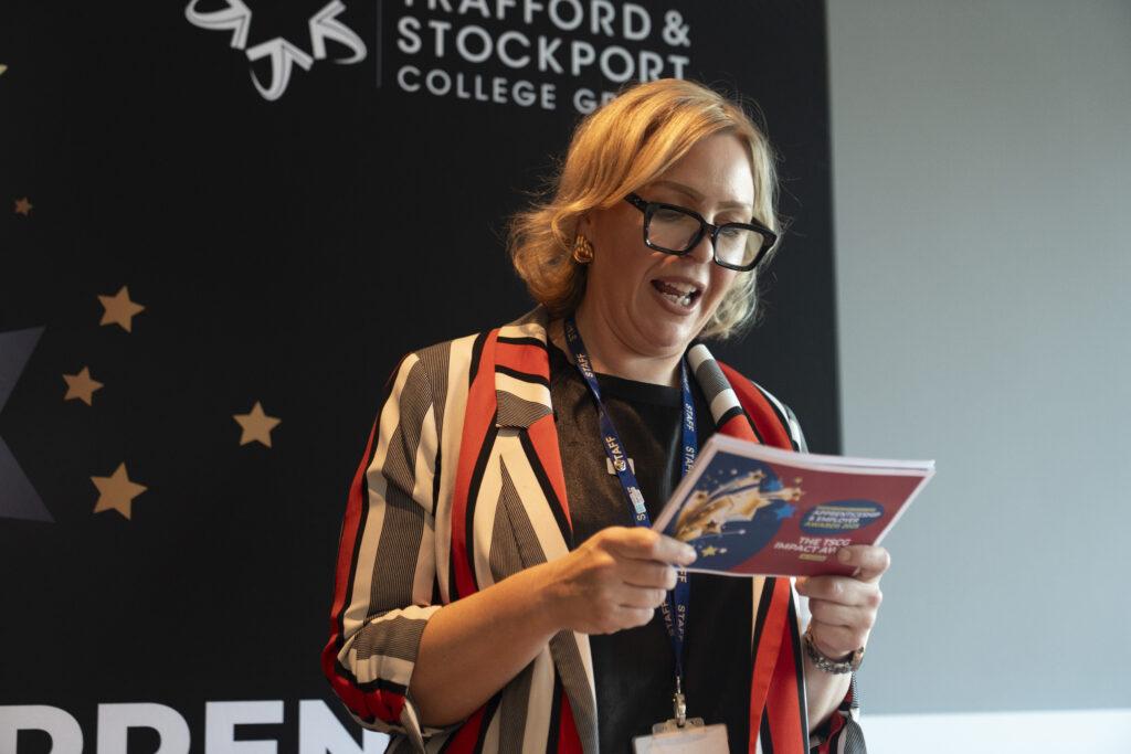 A woman with short blonde hair and glasses, wearing a striped jacket and a "STAFF" lanyard, smiles while reading from an award booklet.