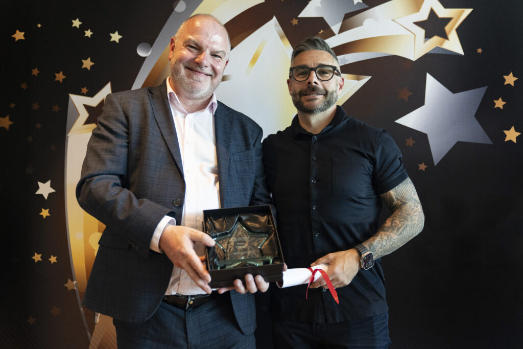 A smiling man in a suit holds a boxed award, standing next to a smiling man with a beard and glasses in a black shirt, who holds a rolled-up certificate. They are against a star-patterned background.