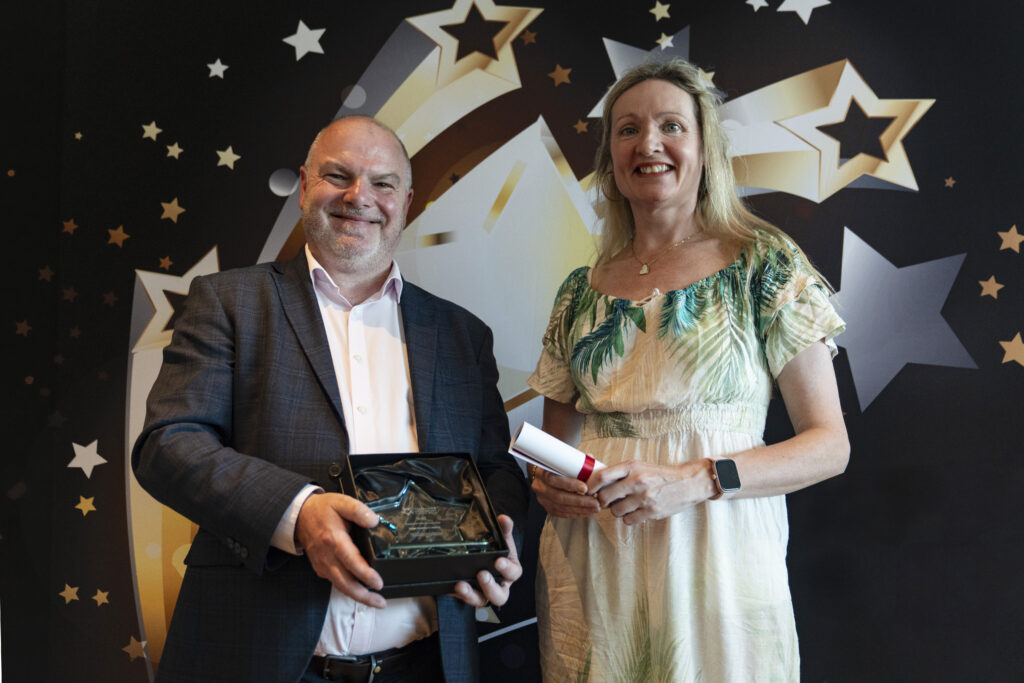  A smiling man in a suit holding an award and a smiling woman holding a certificate, against a star-patterned background.
