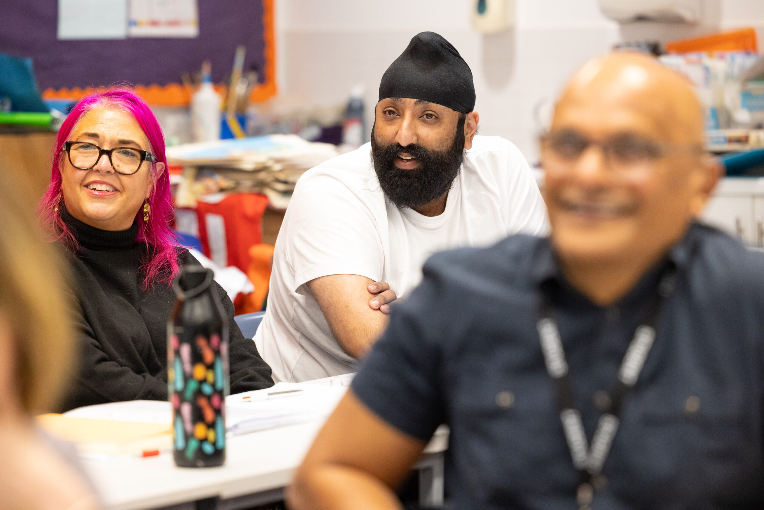 People seated at a classroom table during a workshop or training session, with notebooks, pens, and a patterned water bottle visible on the table. Background shows shelves with supplies and a colourful bulletin board.